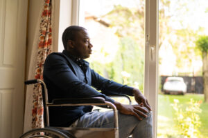 A man is staring dejectedly from his seat in a wheelchair towards a bright sunny day out through a window